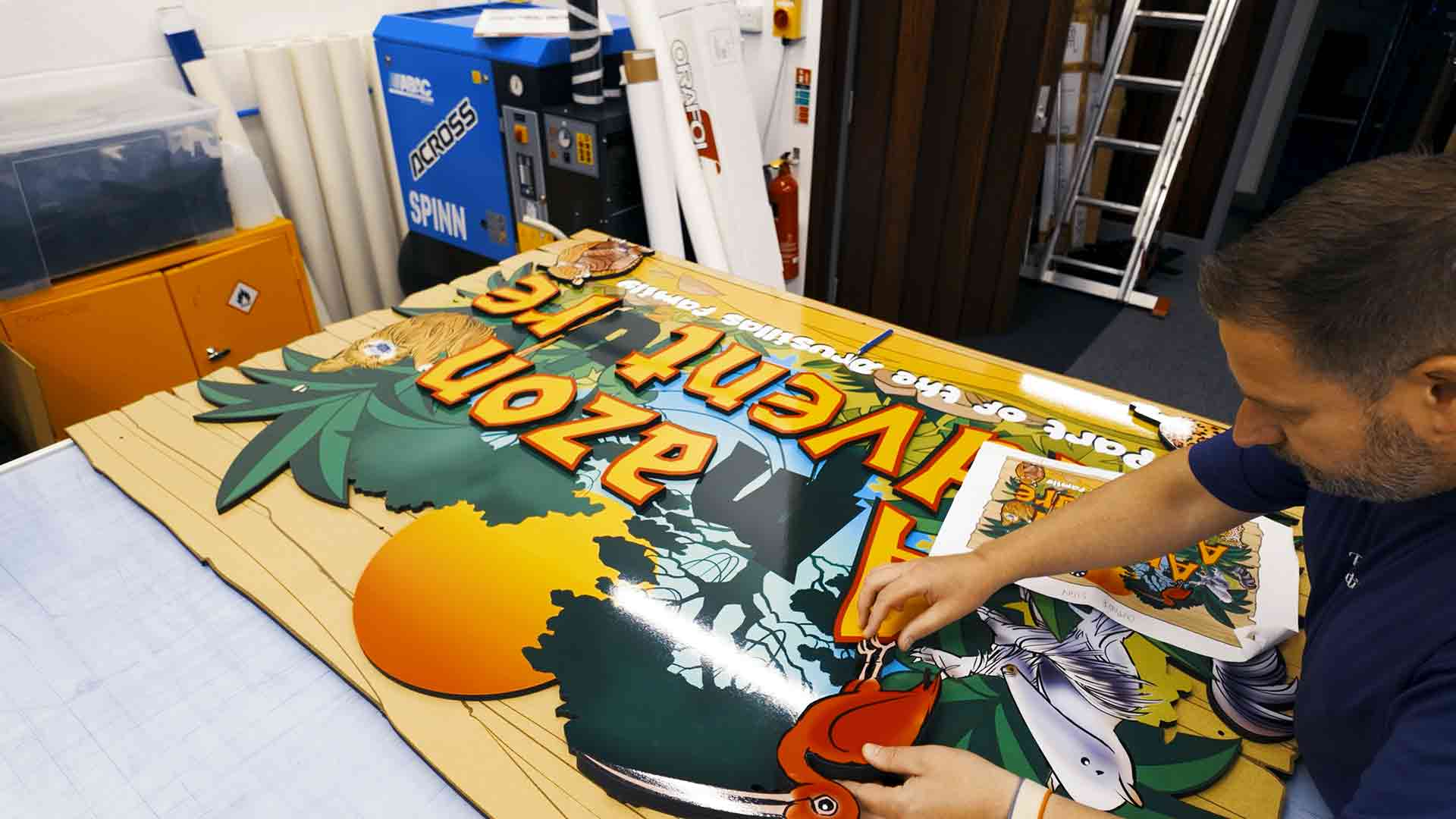 Technician applying finishing touches to the printed “Amazon Adventure” sign for Drusillas Park in the production studio.